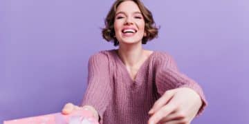 Emotional girl with pretty smile holding pink gift on purple background. Stunning short-haired female model in sweater posing in studio with present..
